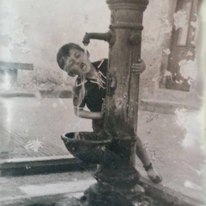 Una fotografia storica in bianco e nero che ritrae un bambino piccolo mentre beve da una fontanella d'acqua in metallo.