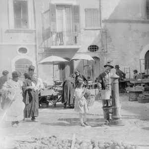 Una fotografia d'epoca in bianco e nero che mostra una scena di vita quotidiana in una piazza di un paese, probabilmente sede di un mercato.