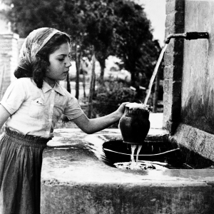 Un ritratto d'epoca in bianco e nero di una giovane ragazza presso una fontana o lavatoio in pietra.