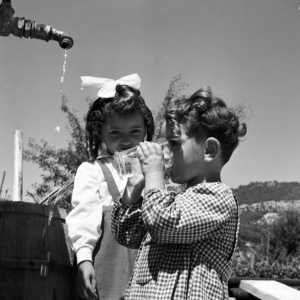 Un primo piano fotografico in bianco e nero che ritrae due bambini piccoli all'aperto vicini a una fonte d'acqua.