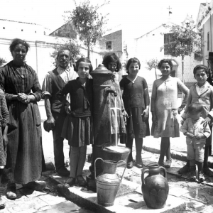 Una fotografia storica in bianco e nero che ritrae un gruppo familiare o di abitanti posizionati attorno a una fontana pubblica "Acquedotto Pugliese".