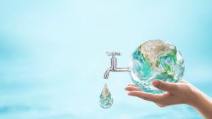 Immagine concettuale che mostra due mani che sorreggono con cura un globo terrestre trasparente. Da un lato del globo emerge un rubinetto cromato da cui cade una singola goccia d'acqua, la quale contiene al suo interno un riflesso della Terra. Lo sfondo è azzurro chiaro e sfocato.